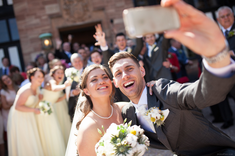 A young bride and groom take a selfie in front of their wedding guests in teh summer sun at Inglewood Manor
